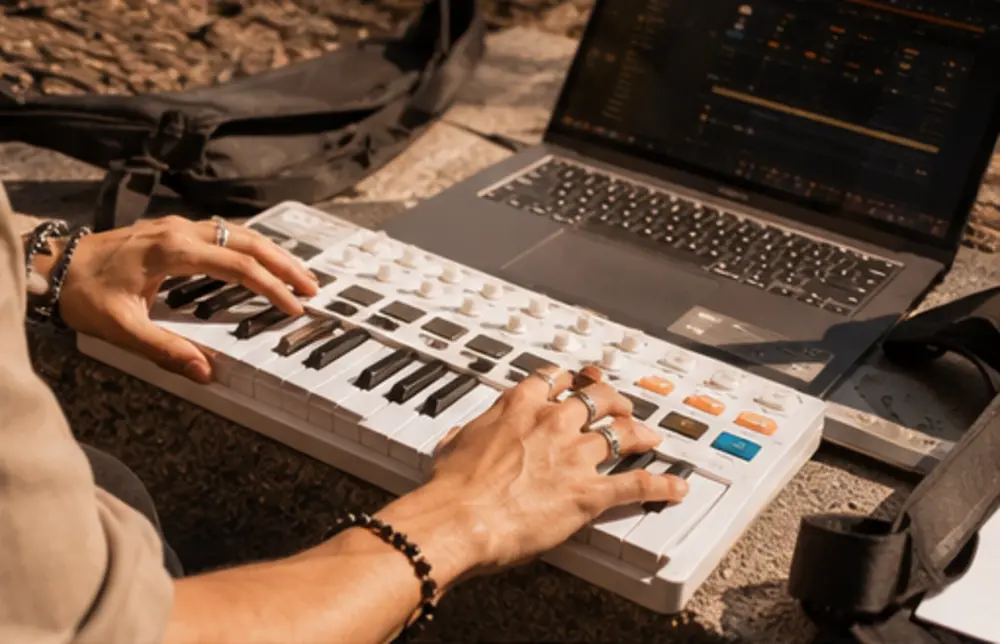 Manos tocando un teclado MIDI junto a una laptop al aire libre